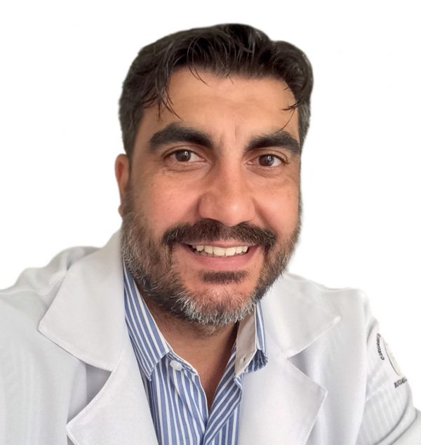Dr. Gustavo Souza de Oliveira - Cirurgião Dentista - está de jaleco branco com uma camisa azul clara listrada por baixo. Ele tem cabelos pretos, barba e bigode.